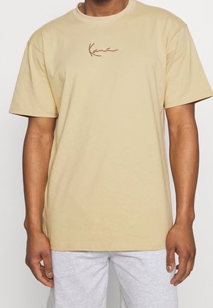 Mężczyzna ubrany w beżowy t-shirt z krótkim rękawem, z małym brązowym logo w stylu kursywy na klatce piersiowej, oraz jasnoszare spodnie, stojący na tle jednolitego tła.