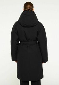 Svart puffjacka med stor huva, bälte i midjan och långa ärmar. Slät textur och knäkort design. Kännetecknas av sin minimalistiska stil.