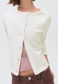 Femeie îmbrăcată cu un cardigan crem descheiat peste un top roz deschis și pantaloni maro, scurtat la umeri și mijloc.