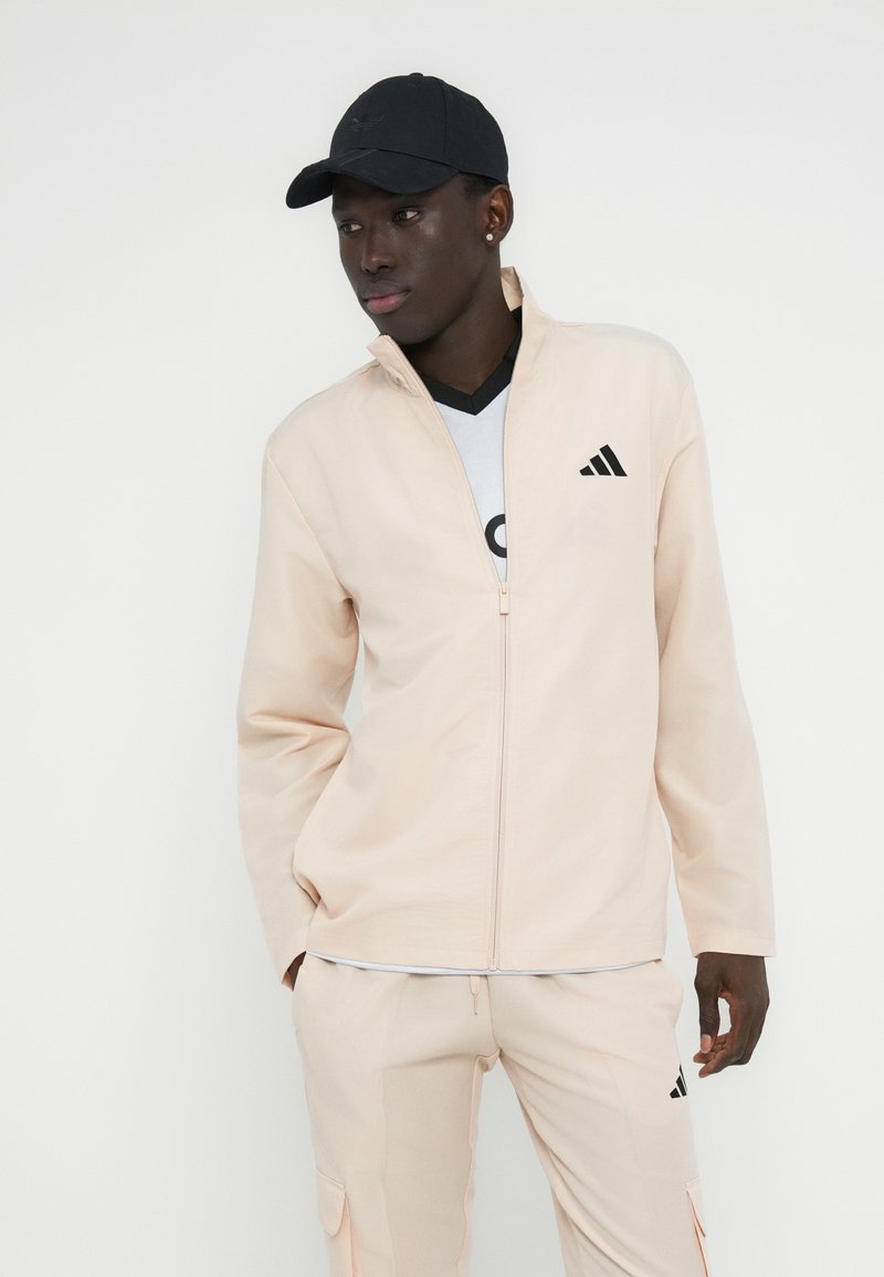 Veste à fermeture éclair couleur pêche claire en tissu lisse, avec un logo adidas sur la poitrine gauche. Associée à un pantalon assorti et une casquette noire.