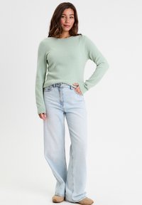 Lys grøn tekstureret sweater med lange ærmer, parret med lys blå brede jeans og beige slip-on fodtøj. Positioneret mod en ensfarvet baggrund.