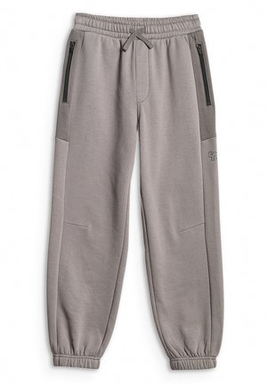 Chłopiec stoi, ubrany w czerwony T-shirt z grafiką samochodu wyścigowego, szare spodnie dresowe, białe skarpetki oraz szare sneakersy z różowymi sznurowadłami na jednolitym tle.