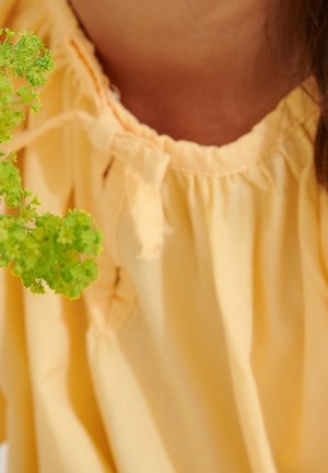 Lichtgele blouse van textuurstof, met een gathered halslijn en strikkoorden. Een groene kruid is gedeeltelijk zichtbaar in de linkervoorgrond.