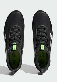 Chaussures de sport noires avec une tige texturée, présentant des rayures blanches, des accents vert fluo et une semelle intérieure imprimée indiquant "METTRE FIN AUX DÉCHETS PLASTIQUES".