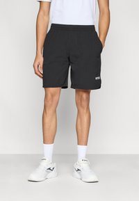 Črne športne kratke hlače z elastičnim pasom, stranskimi žepi in belim logotipom. Oblečene so z belimi športnimi čevlji in nogavicami.