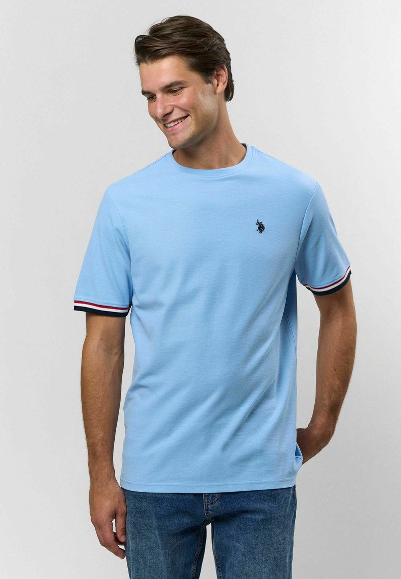 Mladý muž sa usmieva, oblečený v svetlomodrej košeli s krátkym rukávom, s pruhovanými okrajmi rukávov a modrými džínsami, stojí pred jednoduchým pozadím.