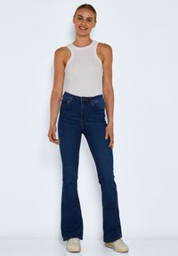 Dark blue flared jeans with a high waist and front pockets, paired with a white ribbed tank top and light-colored sneakers.