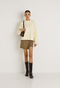 Υπερμεγέθη κρεμ πουλόβερ, μανσέτες με ρίγες, ριγέ φούστα, μαύρες μπότες μέχρι το γόνατο, κρατώντας μικρή καφέ τσάντα, στέκεται μπροστά σε απλό φόντο.