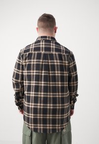 Homme de dos portant une chemise à manches longues à carreaux noir et beige et un pantalon cargo vert olive sur un fond blanc uni.