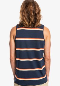 Personne avec des cheveux ondulés aux épaules portant un débardeur bleu marine sans manches avec des rayures horizontales orange et beige, de dos.