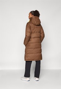 Abrigo acolchado marrón con capucha, presenta secciones acolchadas horizontales, diseño hasta la rodilla y un pequeño logo cerca del dobladillo.