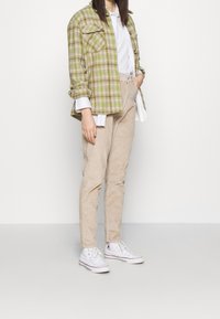 Osoba v béžových kordových kalhotách, bílých vysokých teniskách, bílém tričku a zelené kostkované bundě na prostém bílém pozadí.