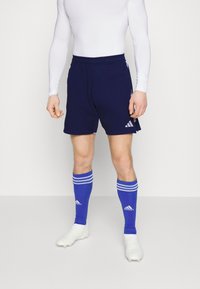 Marineblauwe shorts met het witte Adidas-logo, gecombineerd met blauw gestreepte sokken en witte voetbalschoenen. De outfit heeft een aansluitend wit lange mouw shirt.