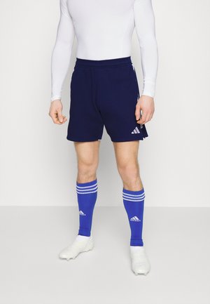 Marineblauwe shorts met het witte Adidas-logo, gecombineerd met blauw gestreepte sokken en witte voetbalschoenen. De outfit heeft een aansluitend wit lange mouw shirt.