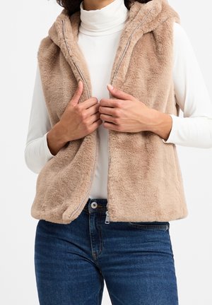 Persona che indossa un gilet beige in ecopelliccia con zip sopra un dolcevita bianco e jeans blu, mani che sistemano il gilet aperto, sfondo neutro.