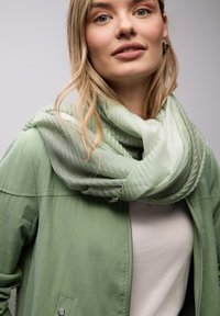 Écharpe vert clair avec un design texturé et superposé, portée sur un haut léger avec une veste verte ; tissu doux et fluide avec un léger éclat.