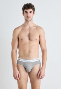 Szürke pamutszálas alsónadrág fehér gumírozott derékkal, melyen a "Calvin Klein" logó található; a modellen rövid, göndör haj van, semleges háttér előtt áll.
