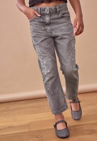 Pantaloni di jeans grigi con dettagli di paillettes luccicanti sulle gambe, a vita alta e taglio cropped. Indossati con scarpe Mary Jane nere glitterate.
