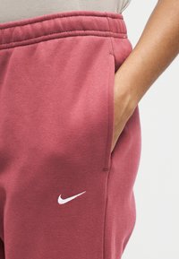 Άτομο φοράει ροζ παντελόνι φόρμας Nike με το χέρι στην τσέπη, εμφανίζοντας το λευκό λογότυπο Nike στο αριστερό μηρό.