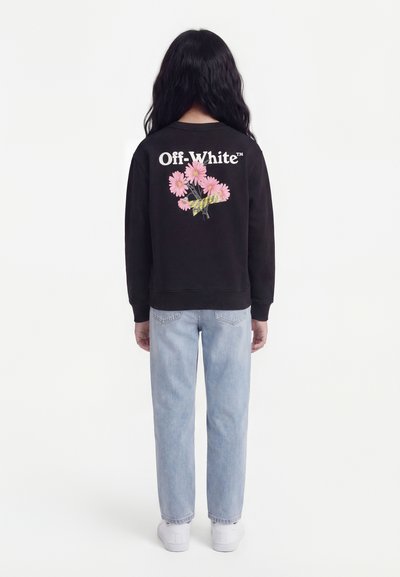 Άτομο με μακριά σκουρόχρωμα μαλλιά στέκεται με την πλάτη, φοράει μαύρο φούτερ με ροζ λουλούδια και το κείμενο "Off-White" στην πλάτη, ανοιχτόχρωμα μπλε τζιν και λευκά παπούτσια.