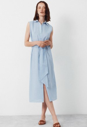 Frau trägt ärmelloses, hellblau-weiß kariertes Midikleid mit Kragen, Taillenband, vorderem Schlitz und braunen Sandalen, steht vor weißer Wand.