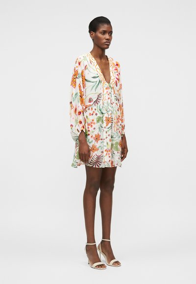 Γυναίκα που φοράει λευκό μίνι φόρεμα με μακριά μανίκια και φλοράλ σχέδιο, με βαθύ V ντεκολτέ και λευκά σανδάλια με ψηλό τακούνι, στέκεται μπροστά σε απλό φόντο.