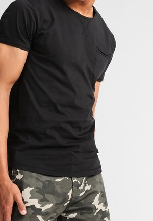 Černé tričko s krátkými rukávy, kulatým výstřihem a přední kapsou, doplněné o maskáčové kalhoty v zelených a béžových tónech.