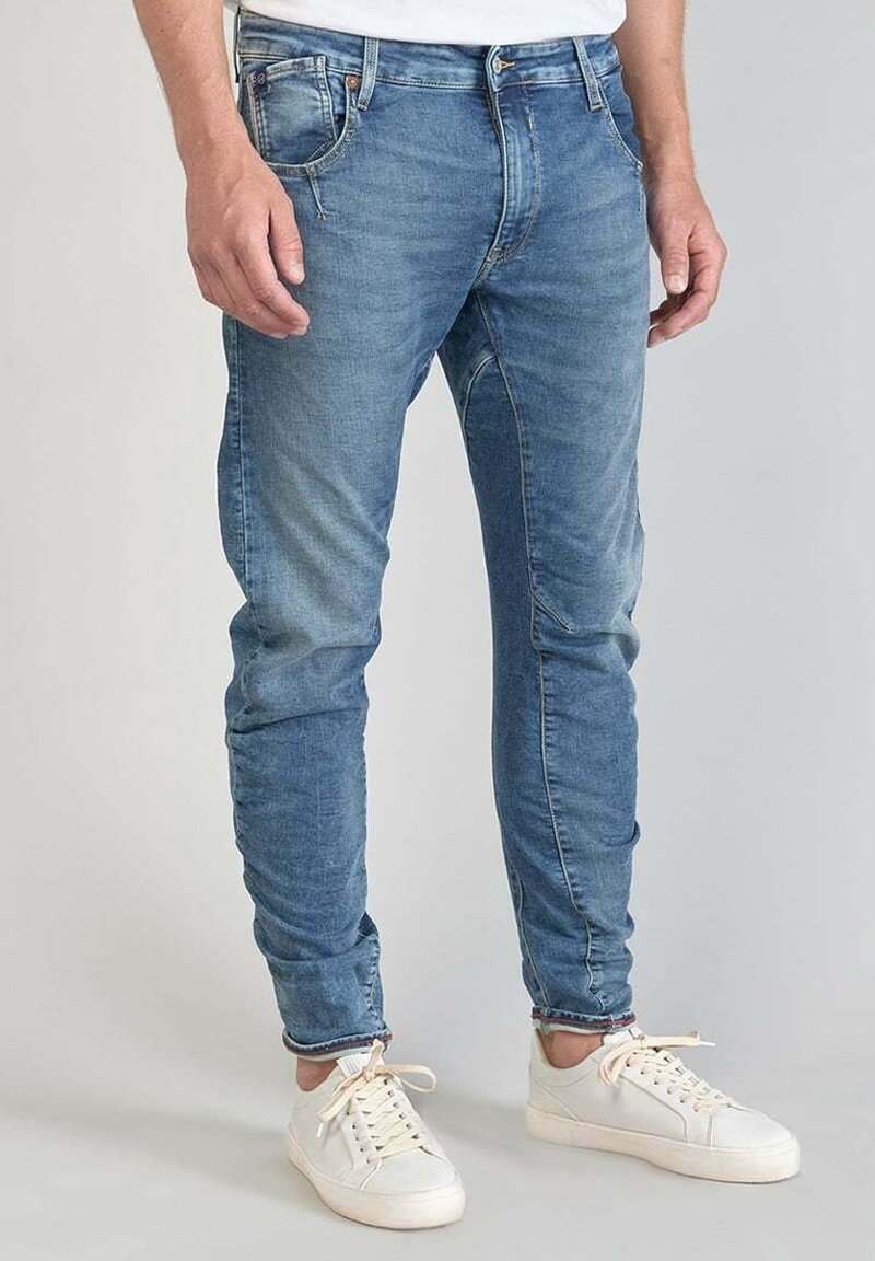 Mężczyzna w niebieskich, obcisłych dżinsach i białych trampkach na jasnoszarym tle, stojący ze swobodnie opuszczonymi rękami.