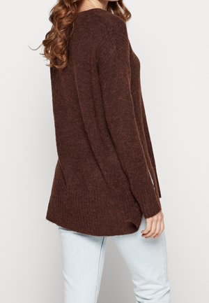 Femme portant un long pull en laine marron avec des fentes latérales et un jean bleu clair, vue de côté et de dos.