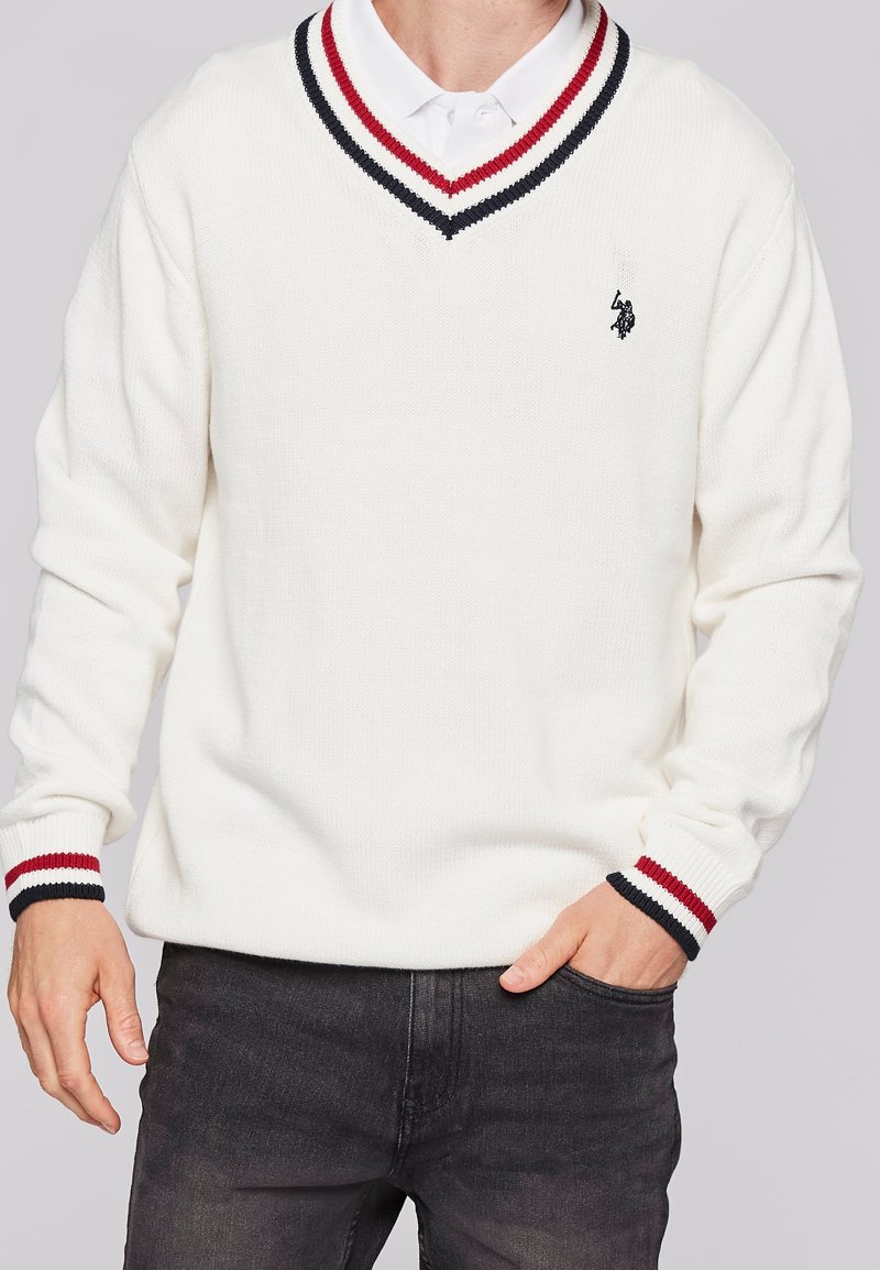 Homme portant un pull blanc à col en V avec des rayures rouges et noires sur le col et les poignets, avec un logo polo sur la poitrine, associé à un jean foncé.