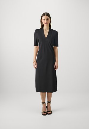 Black, short-sleeve dress with a v-neck and midi length. Smooth fabric with a fitted silhouette. Worn with black heeled sandals and jewelry.