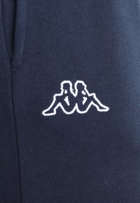 Tums zilas sporta bikses ar mīkstu auduma tekstūru, kurām ir balts izšūts Kappa logo. Dizains ietver elastīgu jostasvietu.