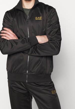 Uomo con le braccia incrociate che indossa una giacca con zip nera EA7 Emporio Armani e pantaloni abbinati con loghi dorati sul petto e sulla coscia.