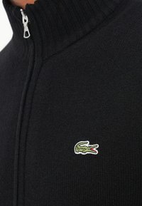 Juoda megzta užtrauktuku palaidinė su raukšlėta apykakle, ant kairio krūtinės puikuojasi mažas žalias ir baltas krokodilo logotipas.