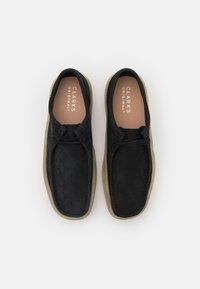 Černé semišové volnočasové boty s zaobleným tvarem, kontrastními béžovými gumovými podrážkami a šněrovacími detaily vpředu. "CLARKS ORIGINALS" vytištěno uvnitř.