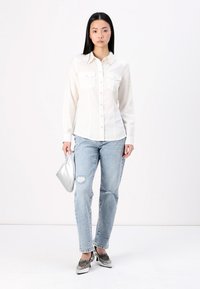 White long-sleeve button-up shirt with chest pockets, paired with light blue distressed jeans and silver flat shoes, holding a metallic clutch.