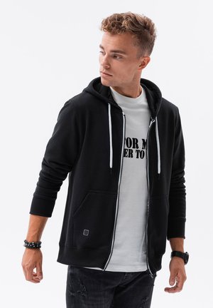 Sudadera con cremallera negra y capucha con cordón, con bolsillos laterales y detalles en blanco, llevada sobre una camiseta blanca con texto en negro.