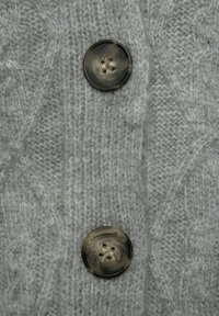 Grijze gebreide trui stof met een gestructureerd visgraatpatroon. Heeft twee grote, gemarmerde knopen met elk vier gaten.
