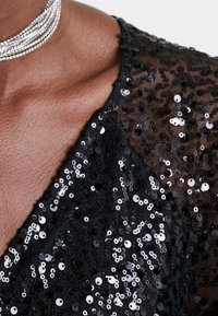 Tissu à paillettes noires avec une surface texturée, présentant un décolleté en V profond. Accompagné d'un collier ras de cou en cristal à plusieurs brins.