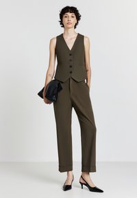 Gilet senza maniche verde oliva con scollatura a V, sei bottoni neri, abbinato a pantaloni su misura e una pochette nera. Eleganti tacchi a punta neri.