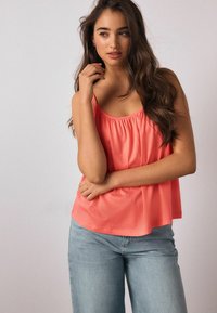 Koralinė palaidinė be rankovių su laisvu siluetu, elastine iškirpte ir plonomis petnešėlėmis, derinama su šviesiai mėlynomis plačiomis džinsų kelnėmis. Minkšta audinio tekstūra.