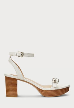 Biała sandałka na wysokim obcasie z drewnianym słupkiem, pasek wokół kostki z klamrą oraz przedni pasek na palce z klamrą na jednolitym tle.