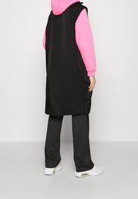 Svart ärmlös väst med sidoknappar, bärs över en rosa hoodie och mörkgrå byxor. Vesten har en slät textur och en lång design.