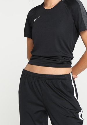 Persoon gekleed in een zwart Nike sportshirt met korte mouwen en zwarte broek met witte zijstrepen, tegen een eenvoudige lichte achtergrond.