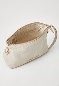 Beige textúrájú bőrtáska cipzárral, rózsaarany kiegészítőkkel és belső zsebbel. A márkanevének megjelenítése az oldalon.