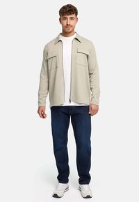Young man wearing beige zip-up shirt jacket, white t-shirt, dark blue jeans, and white sneakers, standing against plain white background.