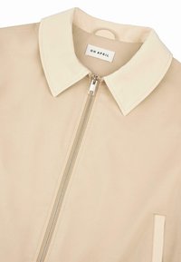 Beige Jacke mit einem kontrastierenden cremefarbenen Kragen und einem Frontreißverschluss. Verfügt über eine Seitenasche und eine glatte Textur. Das Etikett trägt die Aufschrift "OH APRIL."
