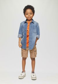 Jeanshemd mit hochgekrempelten Ärmeln über einem orangefarbenen Grafik-T-Shirt, kombiniert mit khakifarbenen Cargo-Shorts und weißen Turnschuhen.