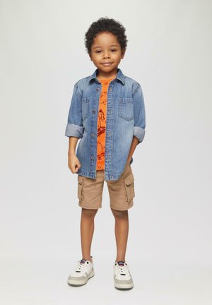 Jeanshemd mit hochgekrempelten Ärmeln über einem orangefarbenen Grafik-T-Shirt, kombiniert mit khakifarbenen Cargo-Shorts und weißen Turnschuhen.