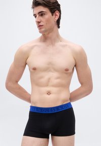 Άνδρας χωρίς πουκάμισο με καστανά μαλλιά, φορώντας μαύρα εσώρουχα Guess με μπλε ζώνη στη μέση, στέκεται με τα χέρια πίσω από την πλάτη, κοιτάζοντας προς τα αριστερά.